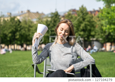 Pleased businesswoman fanning paperwork in hot weather sit in urban public space on lawn enjoy break 127104279