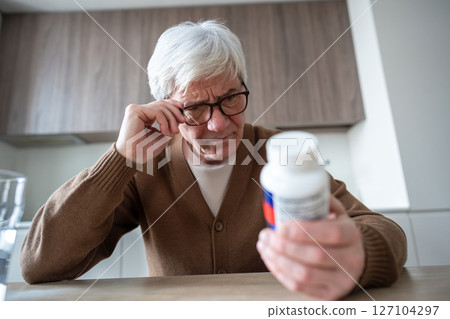 Elderly man squints reading medication label through glasses showing distrust and confusion Elderly man squints reading medication label through glasses showing distrust and confusion 127104297