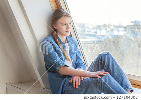 Young happy cute teenage girl in jeans, denim jacket and white T-shirt sitting sitting on the window sill in bright light living room at home and thinking Young happy cute teenage girl in jeans, denim jacket and white T-shirt sitting sitting on the window sill in bright light living room at home and thinking 127104308