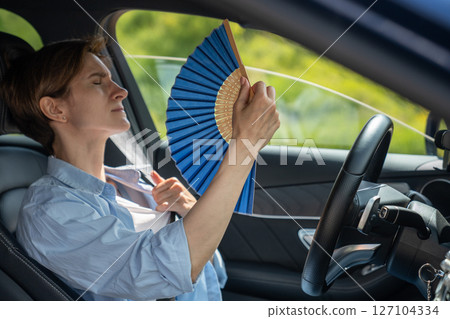 Overheated exhausted middle aged woman driver with hand fan suffering from heat in car. Hot weather  127104334