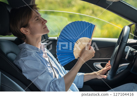 Overheated exhausted middle aged woman driver with hand fan suffering from heat in car. Hot weather  127104416