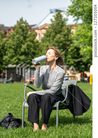 Weary squinting woman drinking water from bottle as rests on grassy lawn in chair in park post work Weary squinting woman drinking water from bottle as rests on grassy lawn in chair in park post work 127105004