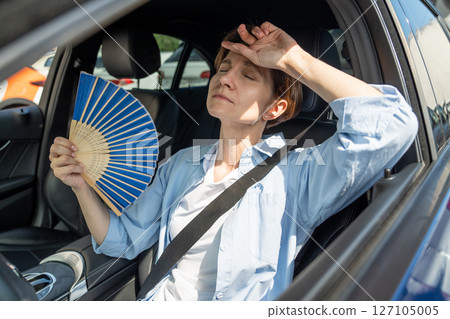 Exhausted middle aged woman drives car waves blue fan suffers from stuffiness in traffic jam. 127105005