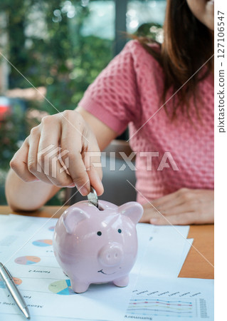 Close-up of a woman placing a coin into a pink piggy bank, surrounded by financial documents, symbolizing savings and financial planning. Close-up of a woman placing a coin into a pink piggy bank, surrounded by financial documents, symbolizing savings and financial planning. 127106547