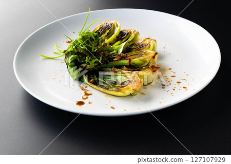 Grilled bok choy garnished with fresh herbs and drizzled with sauce on a sleek black background. 127107929
