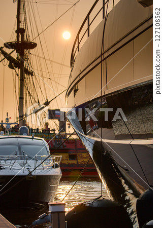 A yacht club at sunset, yachts and sailboats are moored to the berths, huge fenders hang on mega yachts, thick ropes, reflections in the glossy sides of boats, the glare of sun on water surface 127108562