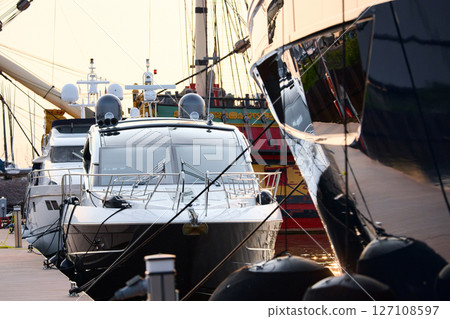 A yacht club at sunset, yachts and sailboats are moored to the berths, huge fenders hang on mega yachts, thick ropes, reflections in the glossy sides of boats, the glare of sun on water surface 127108597
