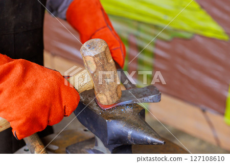Craftsman uses hammer to shape glowing metal on an anvil in axe making setting. 127108610