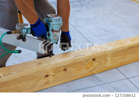 Craftsman focuses on nailing wooden beams together at construction site using pneumatic nail gun 127108612