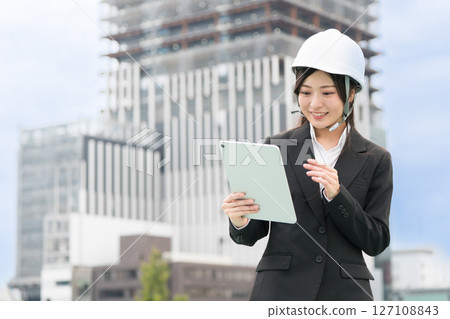 A young woman wearing a suit and helmet operating a tablet; Construction manager, site supervisor, female civil engineer 127108843