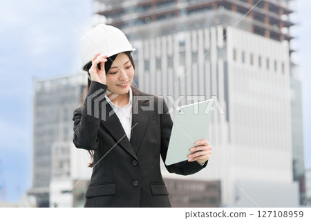 A young woman wearing a suit and helmet checking a tablet; Construction manager, site supervisor, female civil engineer A young woman wearing a suit and helmet checking a tablet; Construction manager, site supervisor, female civil engineer 127108959