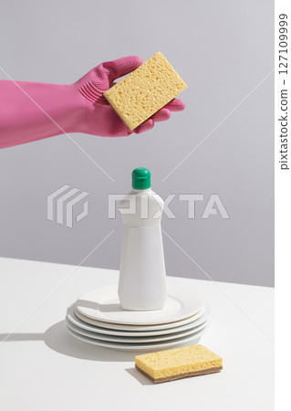 A pink-gloved hand holds a sponge in a dish washing liquid bottle, showing a ready-to-clean setup on a white kitchen or bathroom countertop. Bottle unlabeled for design 127109999