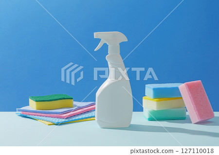 White spray bottle surrounded by soft-colored cleaning sponges, paper, and folded towels, photographed on a crisp, clean blue surface. Concept of cleanliness, cleaning services White spray bottle surrounded by soft-colored cleaning sponges, paper, and folded towels, photographed on a crisp, clean blue surface. Concept of cleanliness, cleaning services 127110018
