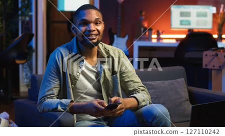 Smiling young adult chatting with friends over texts on mobile phone, sitting on the couch and scrolling social media apps. Young guy staying entertained while working remotely. Camera B. 127110242