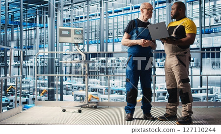 Senior engineer talking with intern in smart factory, collecting and analyzing data. Experienced technician helping trainee use tracking software in industrial plant equipped with sensors, camera A 127110244