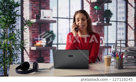 Professional woman in a modern loft takes a phone call at her desk. Multitasking with her laptop, she discusses business strategies, freelance work and career planning. Productivity. Camera B. 127110245