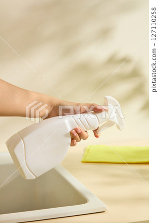 A hand holding a white spray bottle above a kitchen sink, with a yellow cloth nearby. Natural lifestyle imagery of home surface cleaning. Housework, kitchen cleaning 127110258