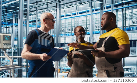 Smart factory engineering team coworkers oversee operation of robotic arms on facility floor. Industry 4.0 plant teamworking colleagues monitor system performance and troubleshoot issues, camera A 127110300