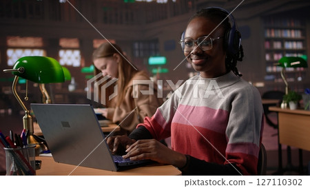 African american girl comparing course notes with data from library collection, using laptop to focus on a literature assignment. Young student working on her diploma project. Camera B. African american girl comparing course notes with data from library collection, using laptop to focus on a literature assignment. Young student working on her diploma project. Camera B. 127110302