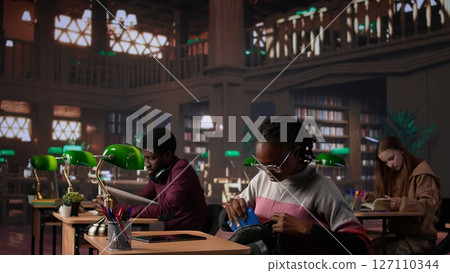 Black girl focuses on her exam preparations in the campus library, takes notes on her laptop from reference books in a peaceful study setting. Girl uses the general database materials. Camera A. 127110344
