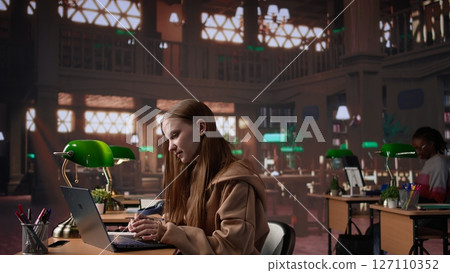 Female student reading and writing detailed notes for an exam, studying political science for upcoming lecture in a library setting at the university. Young adult consults a colleague. Camera B. Female student reading and writing detailed notes for an exam, studying political science for upcoming lecture in a library setting at the university. Young adult consults a colleague. Camera B. 127110352