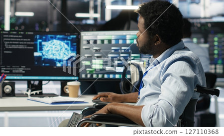 Engineer using wheelchair in data center using AI node tree software to minimize system downtime. Server hub worker in disability using visual programming to monitor performance, camera B Engineer using wheelchair in data center using AI node tree software to minimize system downtime. Server hub worker in disability using visual programming to monitor performance, camera B 127110368