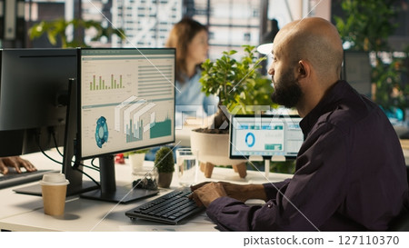 Employee editing documents on desktop computer at office desk, updating reports and files. Worker analyzing data charts on computer screen at desk, interpreting statistical information, camera A 127110370
