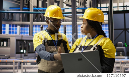 Solar panels factory team working together to fix gear, following maintenance procedures. Photovoltaics plant coworkers doing brainstorming, analyzes data on laptop to predict failures, camera A 127110397