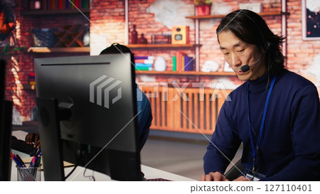 Call center coworkers dealing with upset customers, speaking with them online using headset at office desk. Asian man using PC to do customer support job, handling clients complaints, camera B Call center coworkers dealing with upset customers, speaking with them online using headset at office desk. Asian man using PC to do customer support job, handling clients complaints, camera B 127110401