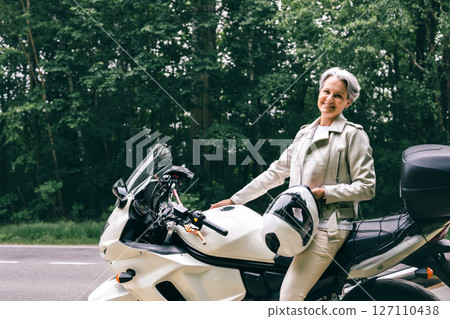 Stylish senior woman with gray hair sitting on motorcycle holding helmet near forest road 127110438