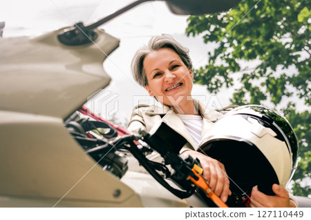 Smiling senior woman with gray hair holding helmet next to motorcycle, confident and free outdoors 127110449
