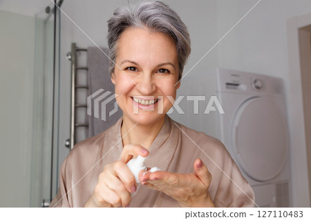 A smiling elderly woman in her age, a pensioner with gray hair, uses a hormonal cream against aging 127110483