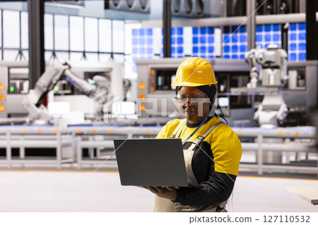 Female engineer in modern facility, overseeing operations and using laptop to ensure efficiency and safety. African american woman in factory, working on production of innovative devices. 127110532