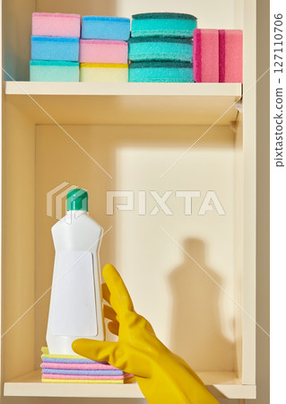 A hand wearing a yellow glove reaches for a bottle of cleaning liquid from inside a shelf filled with stacked multi-colored sponges, revealing a neatly organized cleaning area. A hand wearing a yellow glove reaches for a bottle of cleaning liquid from inside a shelf filled with stacked multi-colored sponges, revealing a neatly organized cleaning area. 127110706