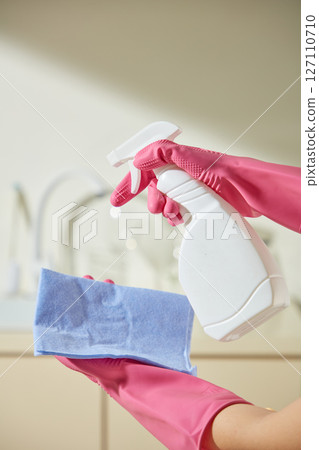 A gloved hand holds a sponge and sprays cleaner onto it in a bright kitchen setting. The image illustrates a cleaning routine with vibrant colors. White spray bottle unlabeled for advertising 127110710