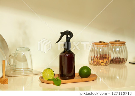 A dark spray bottle sits beside drinking glasses and limes on a light kitchen counter. The scene mixes natural elements with modern cleaning products. 127110713