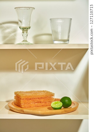 Sponges are stacked next to two glasses on a shelf. Soft lighting highlights a quiet kitchen corner with a clean and cozy atmosphere. advertising cleaning products with green lemon ingredients 127110715