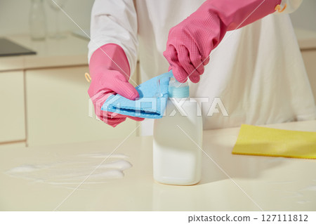 Close-up of a person in pink gloves applying kitchen cleaner to a sponge on a beige countertop, preparing for surface cleaning in a tidy modern kitchen Close-up of a person in pink gloves applying kitchen cleaner to a sponge on a beige countertop, preparing for surface cleaning in a tidy modern kitchen 127111812