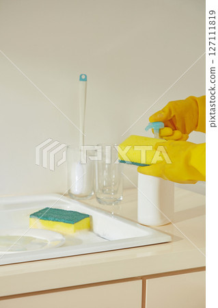 A sponge and pump cleaner placed next to the faucet on a counter, gloved hands preparing for a wipe-down session in a spotless beige kitchen. A sponge and pump cleaner placed next to the faucet on a counter, gloved hands preparing for a wipe-down session in a spotless beige kitchen. 127111819