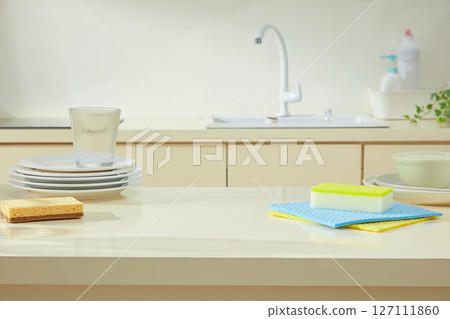 Bright kitchen sink area with stacked dishes, a sponge, and cleaning cloths styled to portray a peaceful yet functional cleaning routine. Kitchen background and blank space for design 127111860