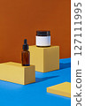 On an orange and blue background, yellow geometric podiums displayed with amber serum bottle and amber jar. Blank space for design label, front view. 127111995