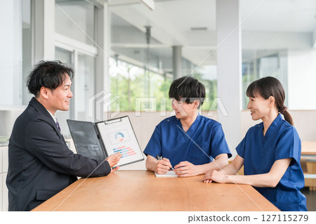 一位來自醫療設備製造商的男性商人和一位穿著手術服的男性和女性醫務人員正在進行銷售和商業談判 127115279