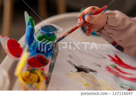 Close up of a toddlers hand reaching for a paintbrush with gouache colors. Creative activity at home, early childhood art education. Close up of a toddlers hand reaching for a paintbrush with gouache colors. Creative activity at home, early childhood art education. 127116047