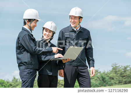 Male and female workers in work clothes having a meeting while looking at a laptop at a construction site 127116290