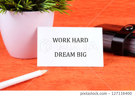 Motivational words WORK HARD DREAM BIG written on a card next to a small potted plant and stationery 127116400