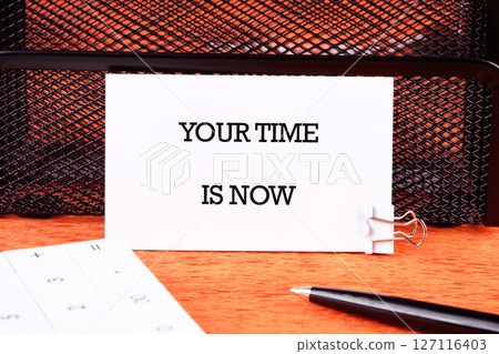 Motivational concept words YOUR TIME IS NOW written on a business card next to a pen and a calculator in a prominent place 127116403
