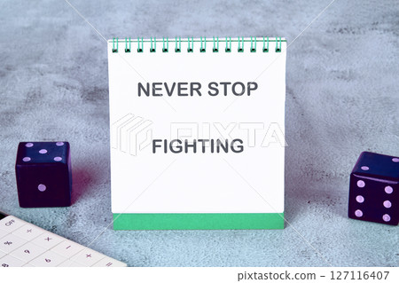 The concept of inspirational and motivational. NEVER STOP FIGHTING written on a desktop notebook, surrounded by a calculator and dice 127116407