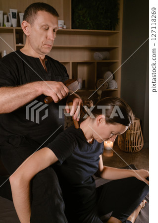 Male master giving Thai Toksen massage to girl in massage room. A girl gets a relaxing Thai back massage with a Toxen massage kit Male master giving Thai Toksen massage to girl in massage room. A girl gets a relaxing Thai back massage with a Toxen massage kit 127118269