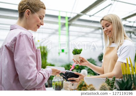 A customer pays for a purchase at a florist shop using a smartphone. The shop assistant is offering a small plant. 127119196