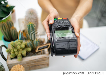 A florist offers a point-of-sale device at a counter with cacti visible in the background, promoting commerce. 127119219